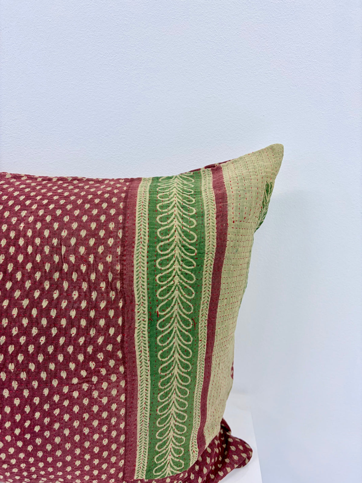 A close-up of a decorative throw pillow featuring a maroon and beige dotted pattern, a vertical green band with cream leaf embroidery, and a beige woven side, set against a plain light blue wall.
