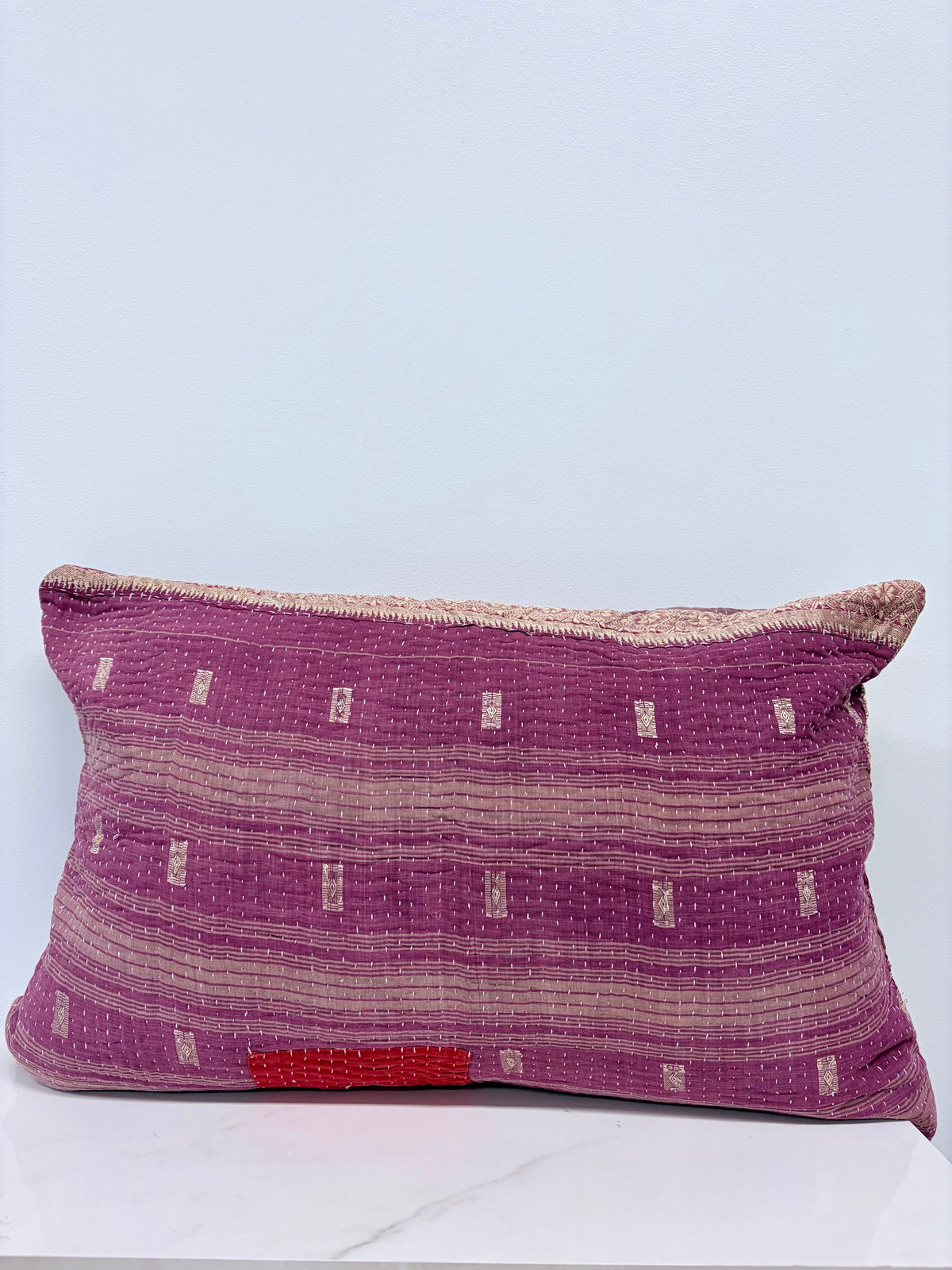 A rectangular throw pillow made from purple and beige woven kantha fabric with a small red accent patch near the bottom, resting on a white marble surface against a light blue-gray background.