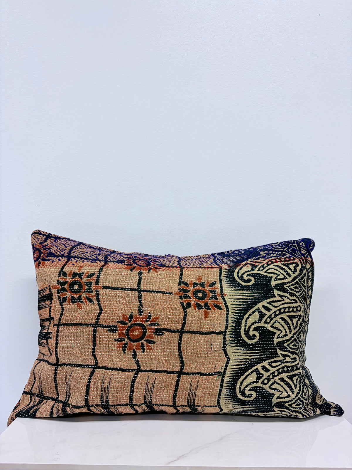 A square decorative throw pillow with a vintage kilim textile pattern in beige, black, and blue, resting on a white marble surface against a light gray wall.