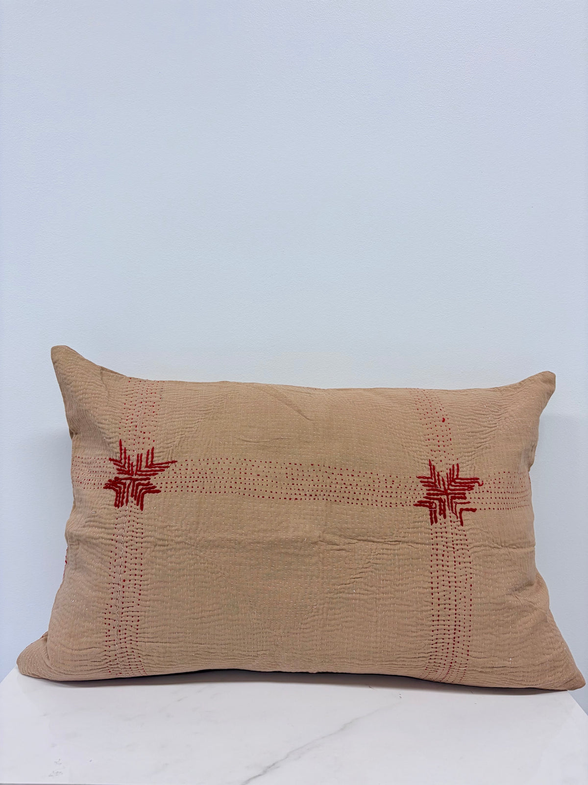 A square beige linen throw pillow with red embroidered star and dotted line patterns, displayed on a white marble surface against a light blue-gray background.