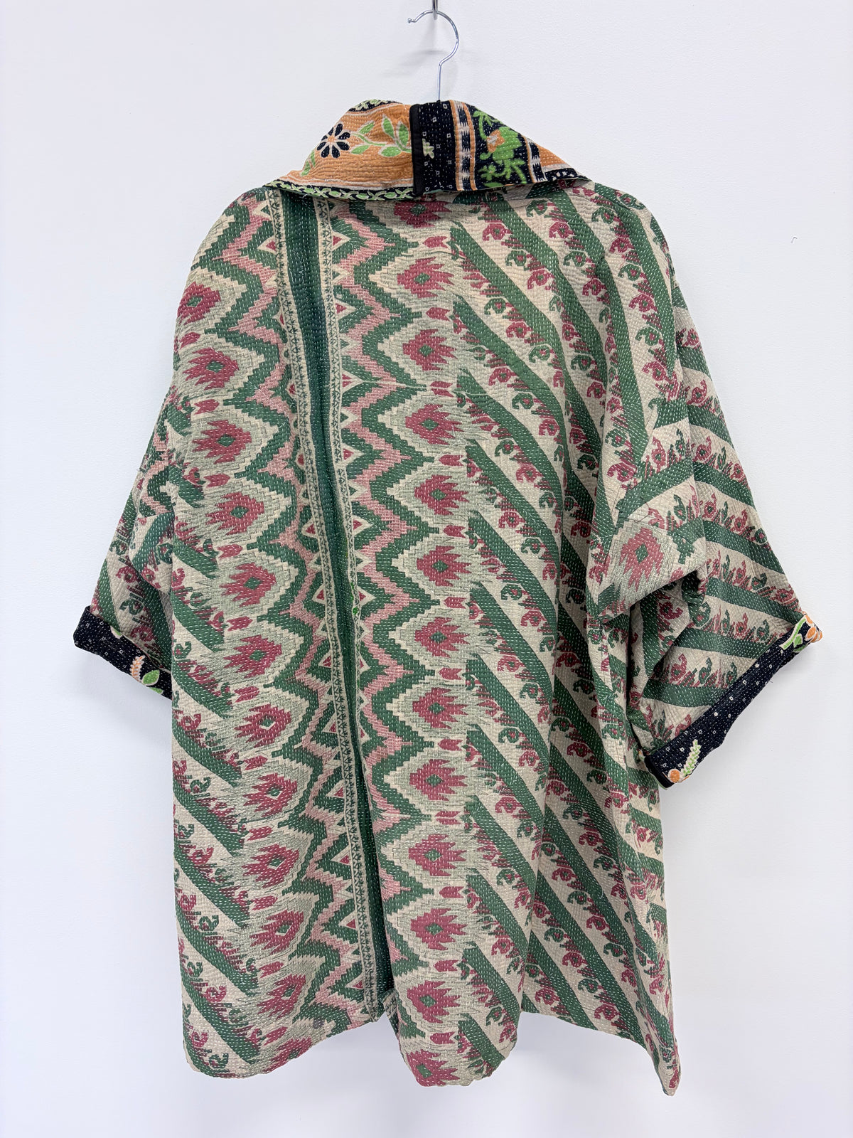 A vintage patterned kaftan-style tunic top hanging on a white hanger against a white background. The garment features a green and beige geometric ikat pattern with pink floral accents, a dark green center panel, and a multicolor patterned collar.