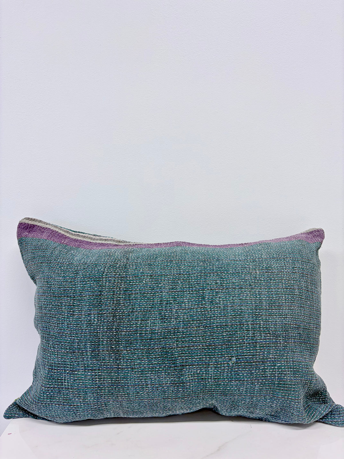 A square woven throw pillow in a textured teal blue fabric with a subtle purple stripe along the top edge, set against a plain light gray background.