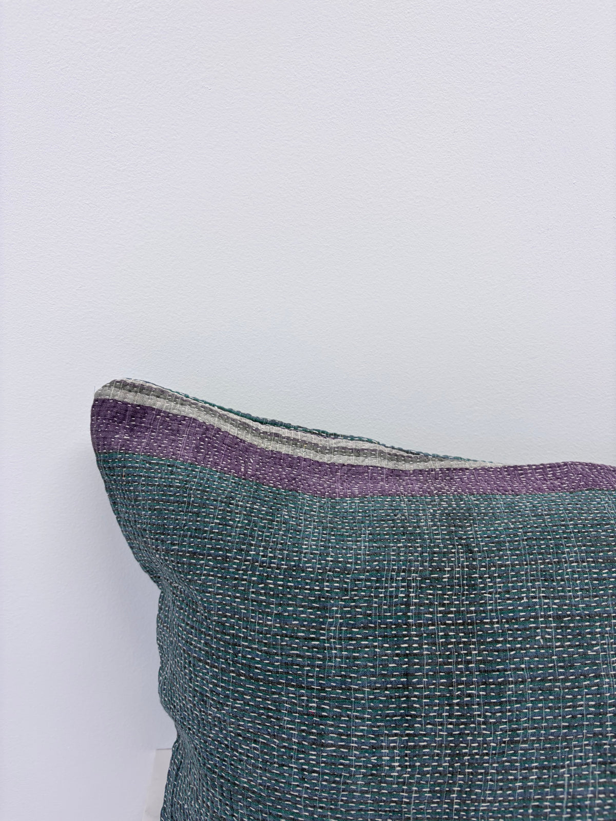 A close-up of a woven throw pillow corner showing a textured teal-green body with a purple stripe and thin gray and white accent stripes along the top edge, set against a plain white wall.