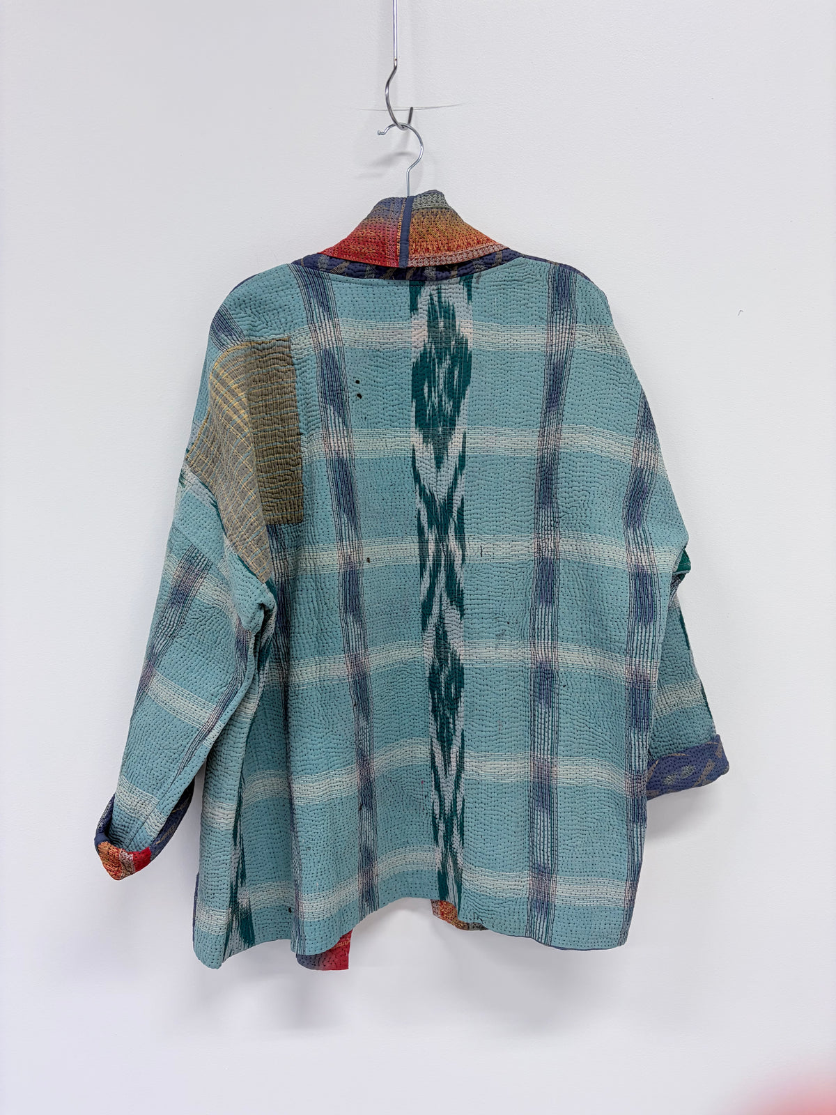 A handmade patchwork long-sleeve shirt in teal blue, gray, and brown plaid with ikat patterns, hanging on a wooden hanger against a white wall.