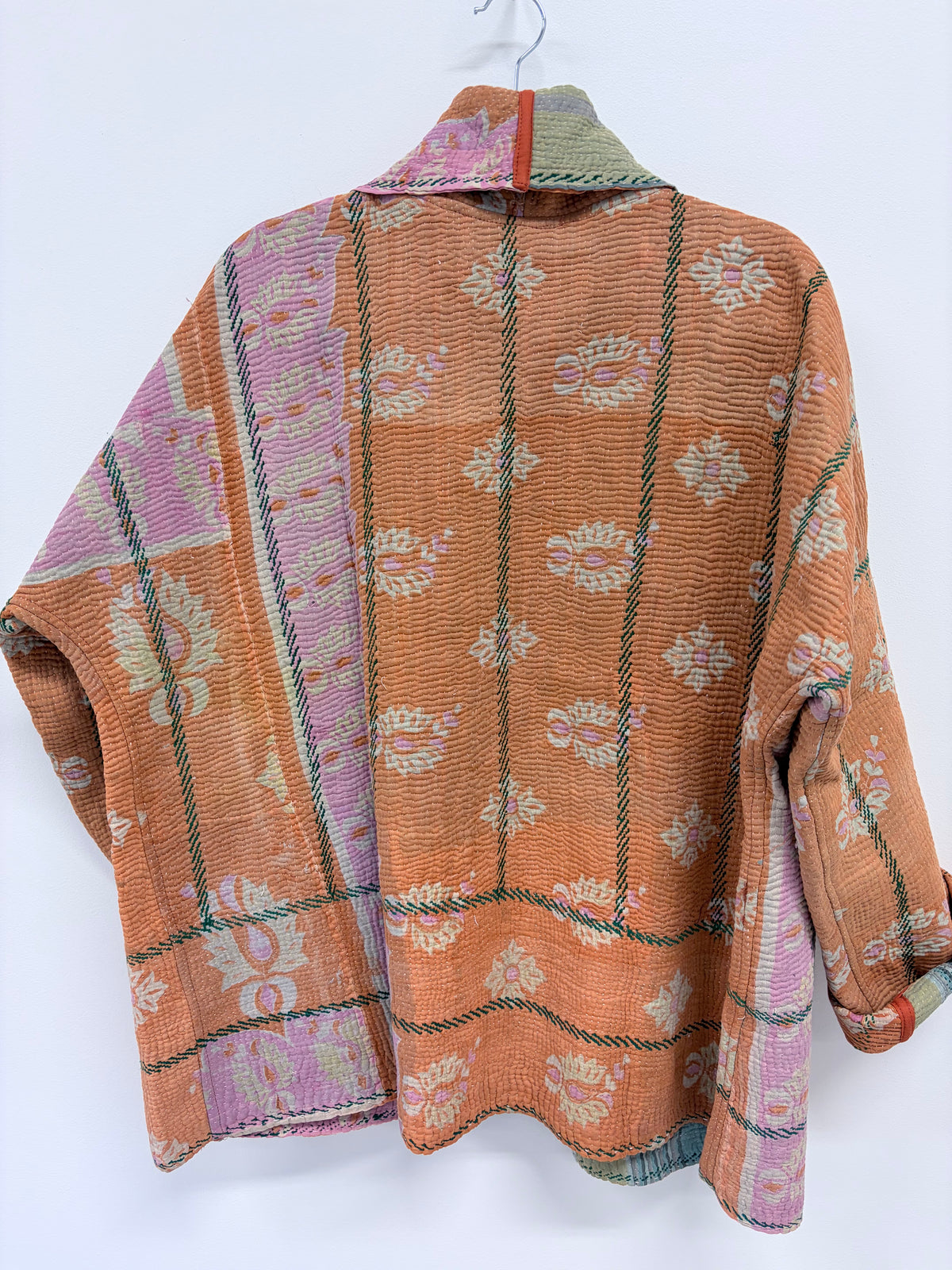 A textured orange cotton shawl collar jacket with pink, beige, and green woven paisley patterns, hanging on a white wall.