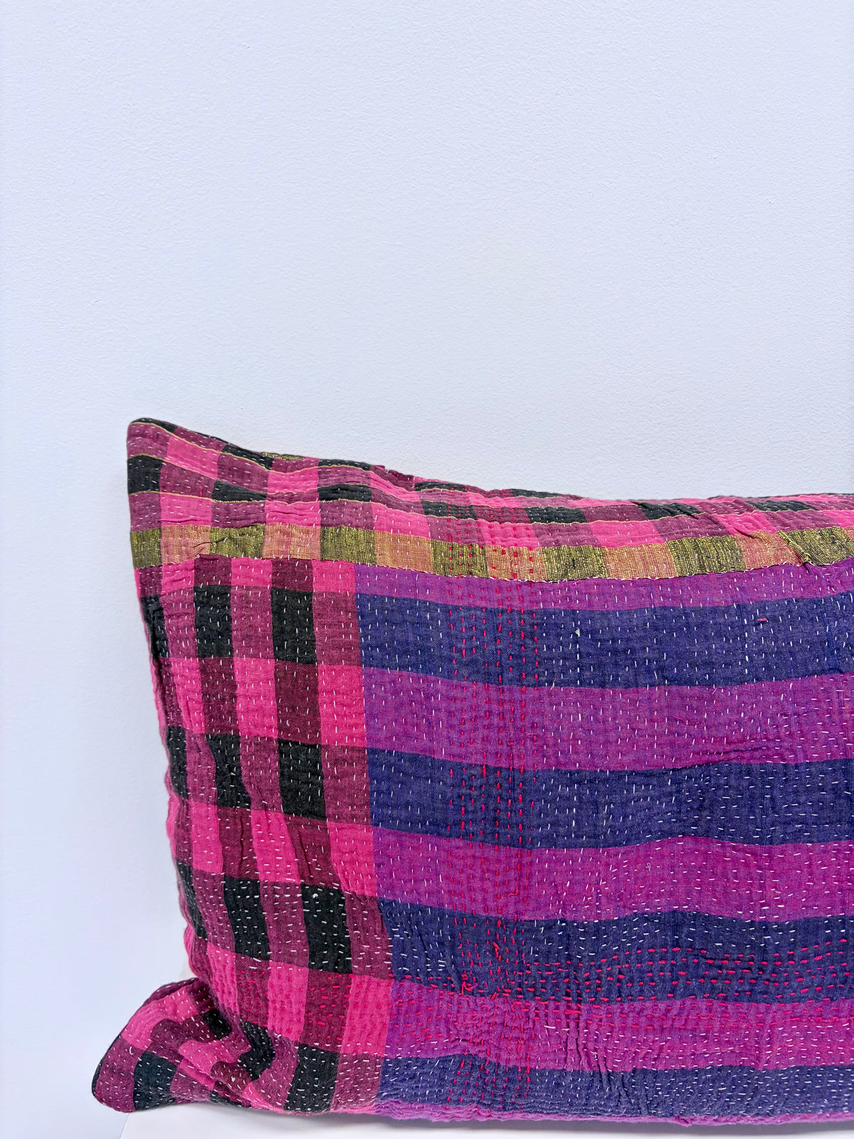 A close-up of a textured patchwork throw pillow featuring vibrant pink, purple, black, and olive green plaid and checkered patterns, set against a plain light gray background.