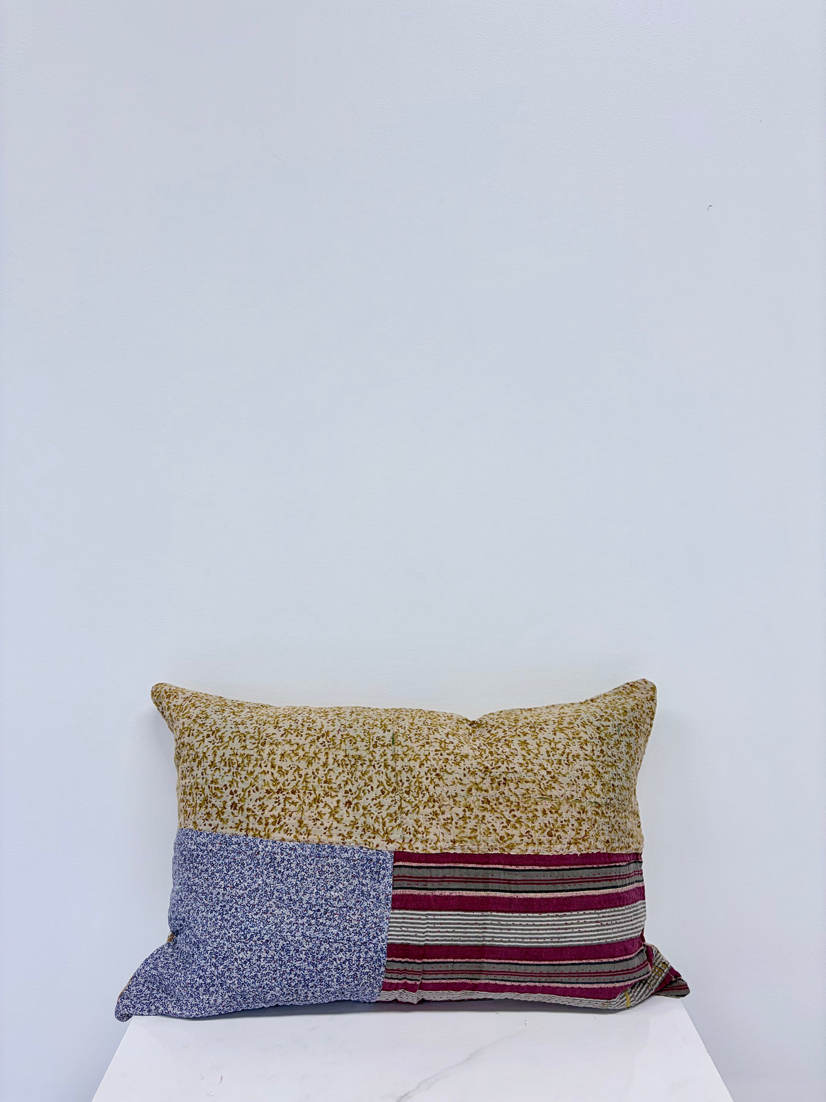 A rectangular patchwork lumbar pillow with three distinct fabric panels, resting on a white marble-topped surface against a plain white wall.