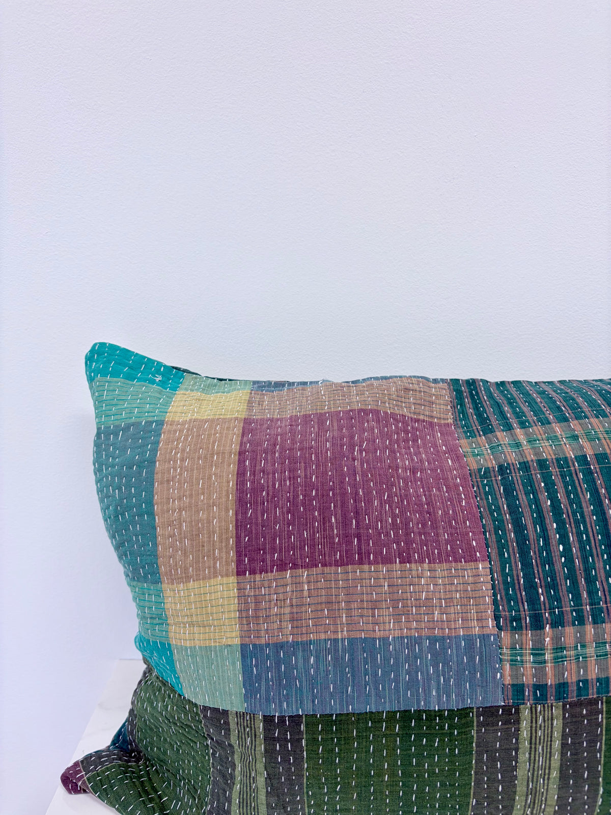 A close-up of a patchwork kantha pillow with a plaid pattern in teal, purple, green, and beige, featuring visible running hand-stitching.
