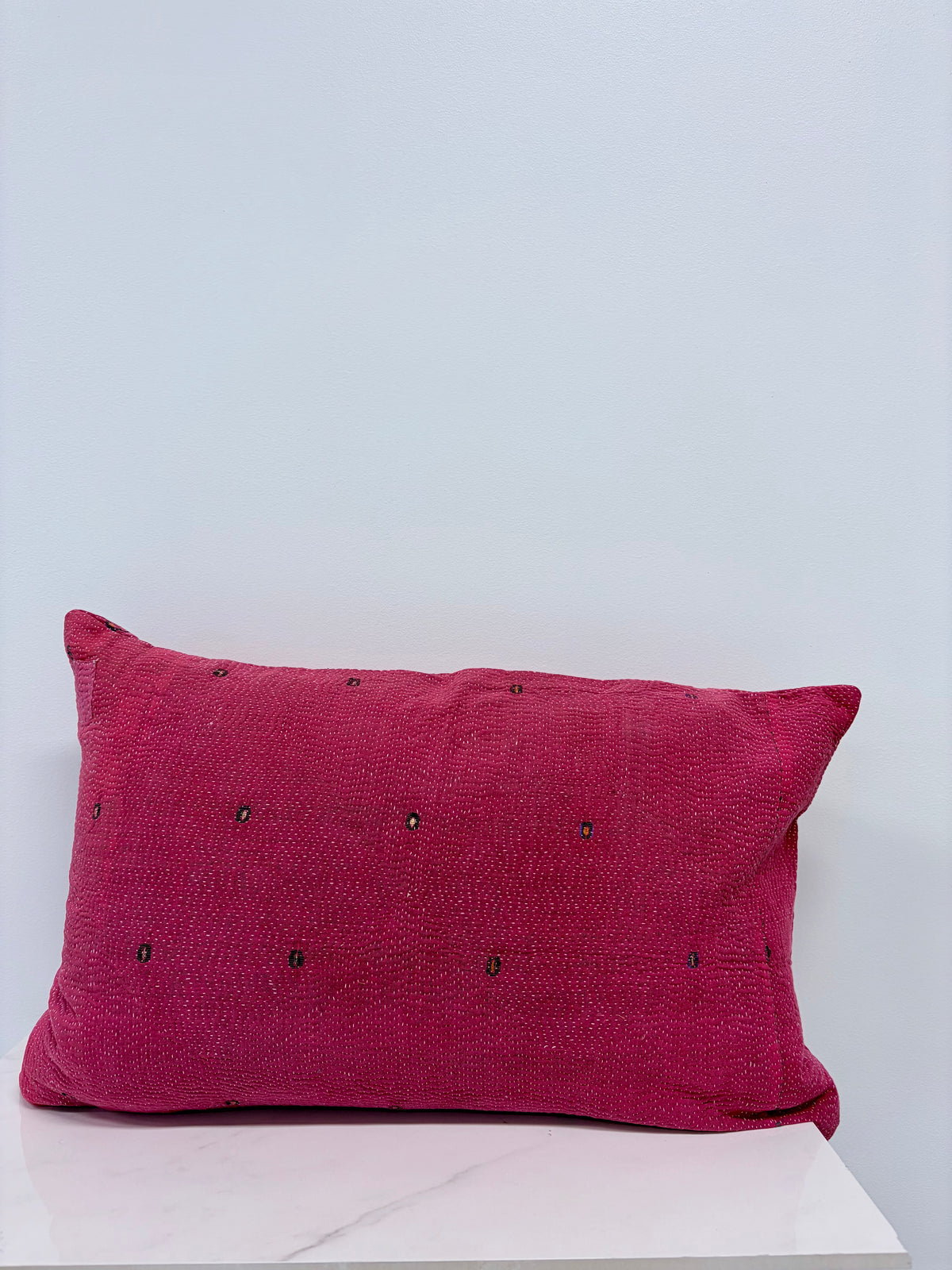 A rectangular magenta throw pillow with small black embroidered dots, resting on a white marble surface against a light gray wall.