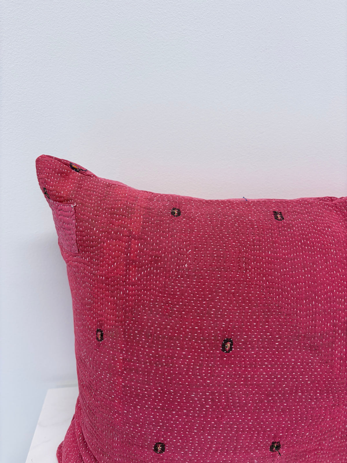 A close-up of a textured, woven pink throw pillow with small black embroidered details, set against a plain white wall.