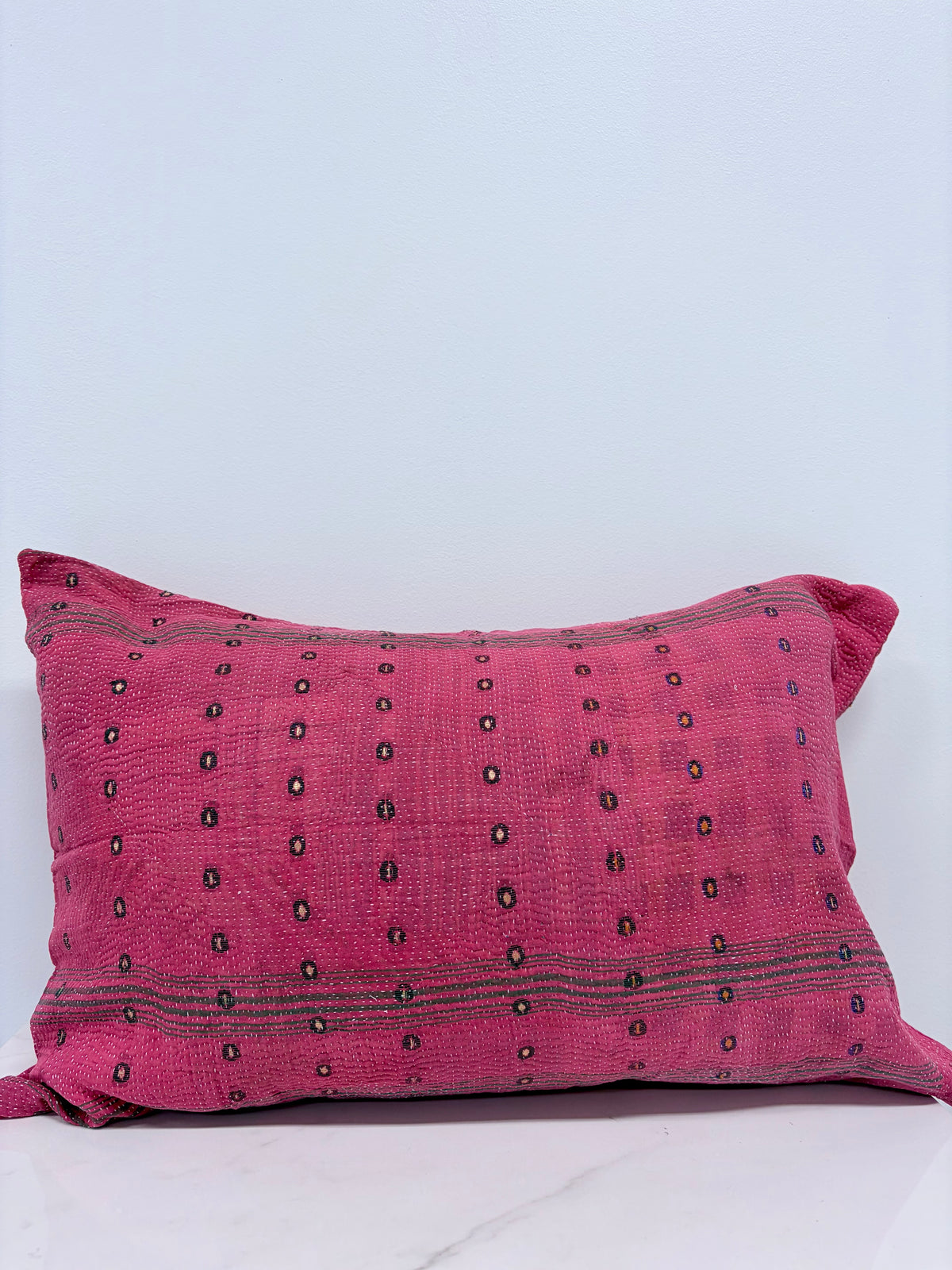 A rectangular pink throw pillow with a textured woven fabric, featuring small black embroidered dots and horizontal stripes, set against a plain light gray background.
