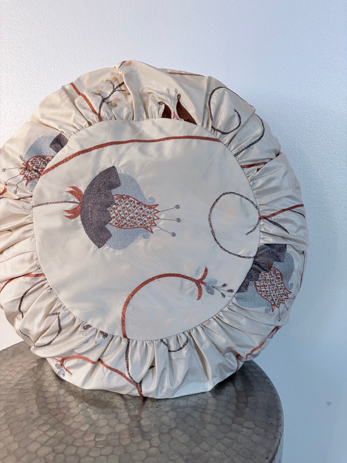 A round decorative pillow with a gathered ruffled edge, featuring a central printed panel with stylized floral motifs in gray, rust red, and black on a cream background, displayed on a metallic table against a light wall.
